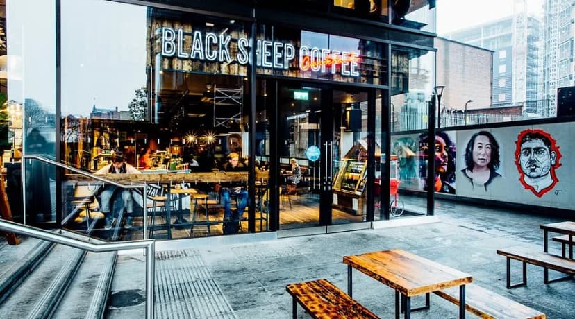 Black Sheep Coffee storefront with full-glass facade, neon signage, outdoor seating and street-art portraits on the adjacent wall