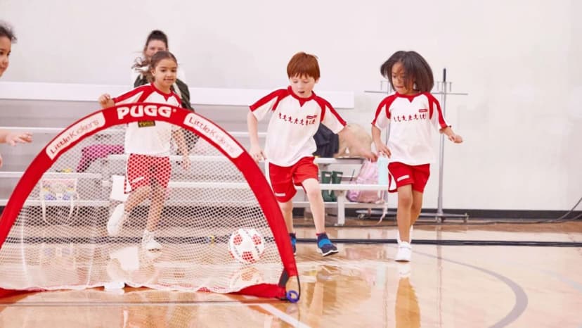 Young children in red and white Little Kickers kit playing a football game indoors with a pop-up PUGG goal in a school sports hall