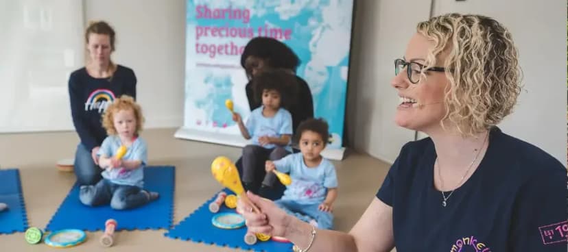 A Monkey Music teacher wearing a headset mic and holding bright-yellow maracas leads a class of mothers and toddlers sitting on blue play mats beneath a 'Sharing precious time together' brand banner