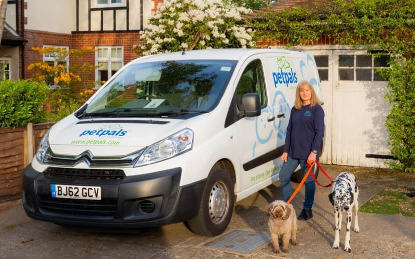 A Petpals franchisee in a branded navy fleece standing beside her white Petpals Citroën van, holding orange leads attached to a brown labradoodle and a black-and-white dalmatian on a suburban driveway