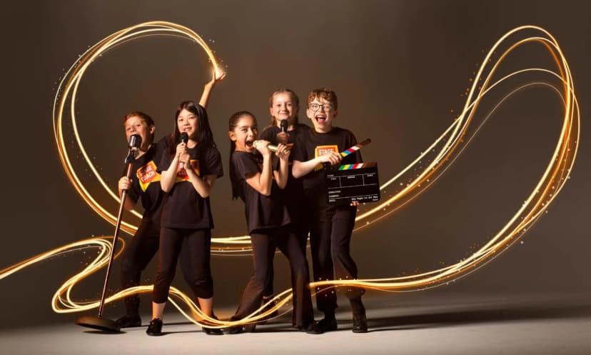Five Stagecoach Performing Arts students in black tees with orange badge logos posing dramatically with microphones and a film clapperboard, surrounded by swirling gold light trails that suggest a sparkling stage performance