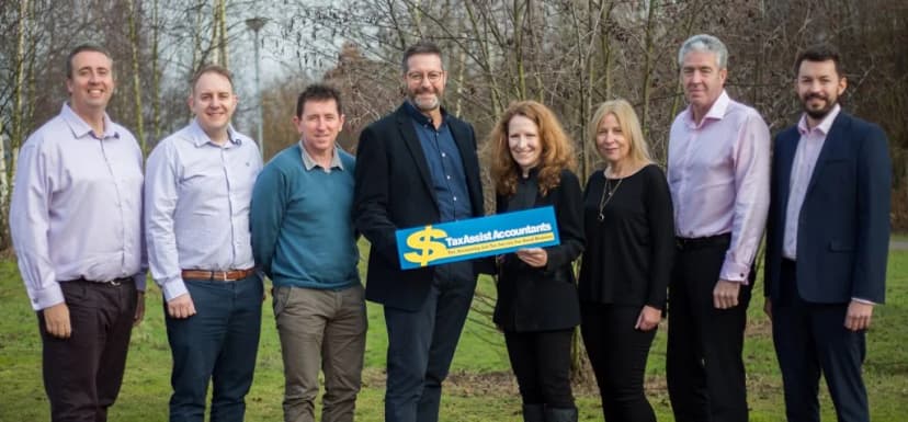 Seven TaxAssist Accountants franchisees and head-office staff standing outdoors in winter holding a TaxAssist branded sign reading 'The Accounting and Tax Service For Small Business' - illustrating the network's people and community