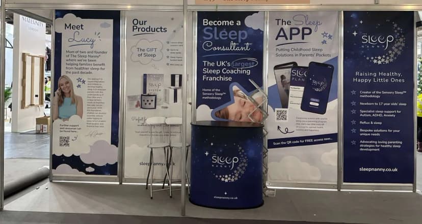 The Sleep Nanny franchise exhibition stand with five branded banner panels - 'Meet Lucy' (founder profile), 'Our Products: The Gift of Sleep', 'Become a Sleep Consultant: Home of the Sensory Sleep methodology and the UK's largest Sleep Coaching Franchise', 'The Sleep APP', and a Sleep Nanny brand panel listing benefits